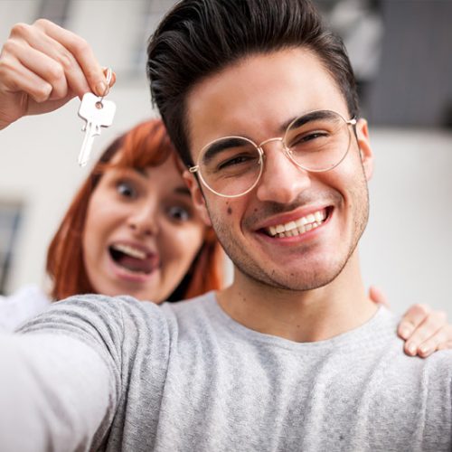 A smiling young man takes a selfie while a woman behind him excitedly holds up keys, suggesting they have just acquired a new home. Both appear happy and celebratory.